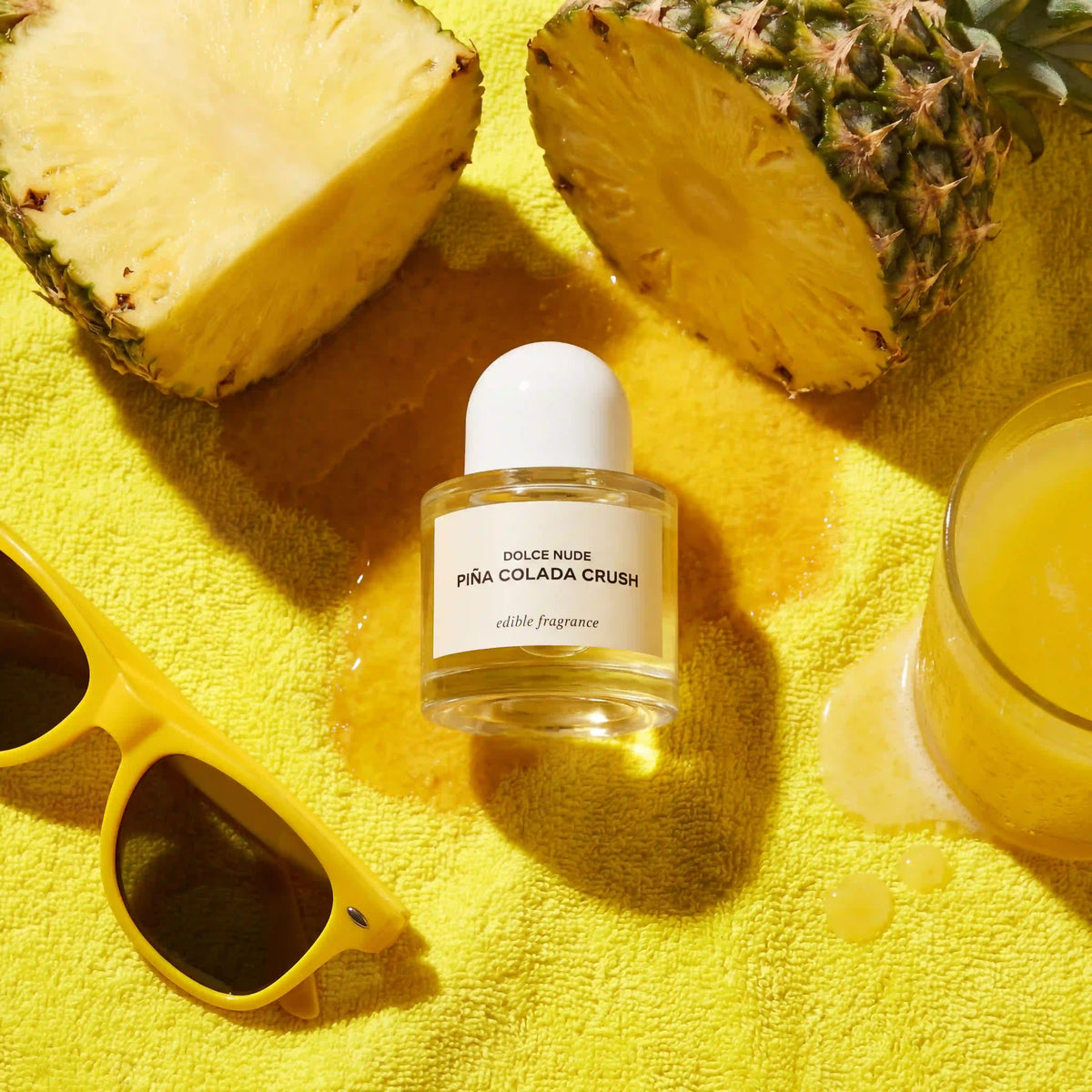 DOLCE NUDE Piña Colada Crush edible fragrance bottle surrounded by fresh pineapple slices and yellow sunglasses on tropical background